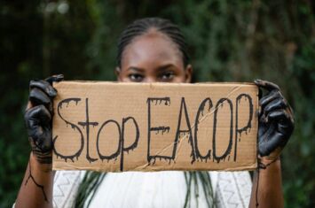 Eine Person mit Erdölbeschmierten Händen hält ein Schild, auf dem steht »Stop EACOP«.