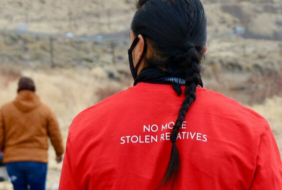 Zwei Personen sind von hinten zu sehen. Auf dem roten Kleidungsstück der Person im Vordergrund ist der Satz "No more stolen relatives" zu lesen. Der schwarze Haarzopf der Person verdeckt Buchstaben der Botschaft. Im Hintergrund ist hügliges Land zu sehen.