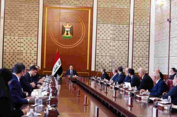 An einem langen Tisch sitzen viele Männer in Anzügen, dem Vorsitzenden, der neben einer irakischen Flagge sitzt, zugewandt