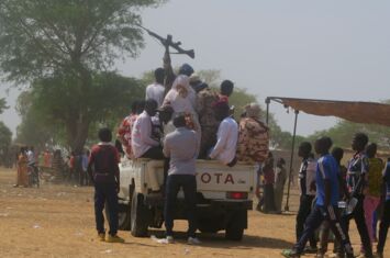 Viele Personen, zum Teil in Uniform, drängen sich auf der Ladefläche eines Transporters, einer hält ein Gewehr in die Höhe