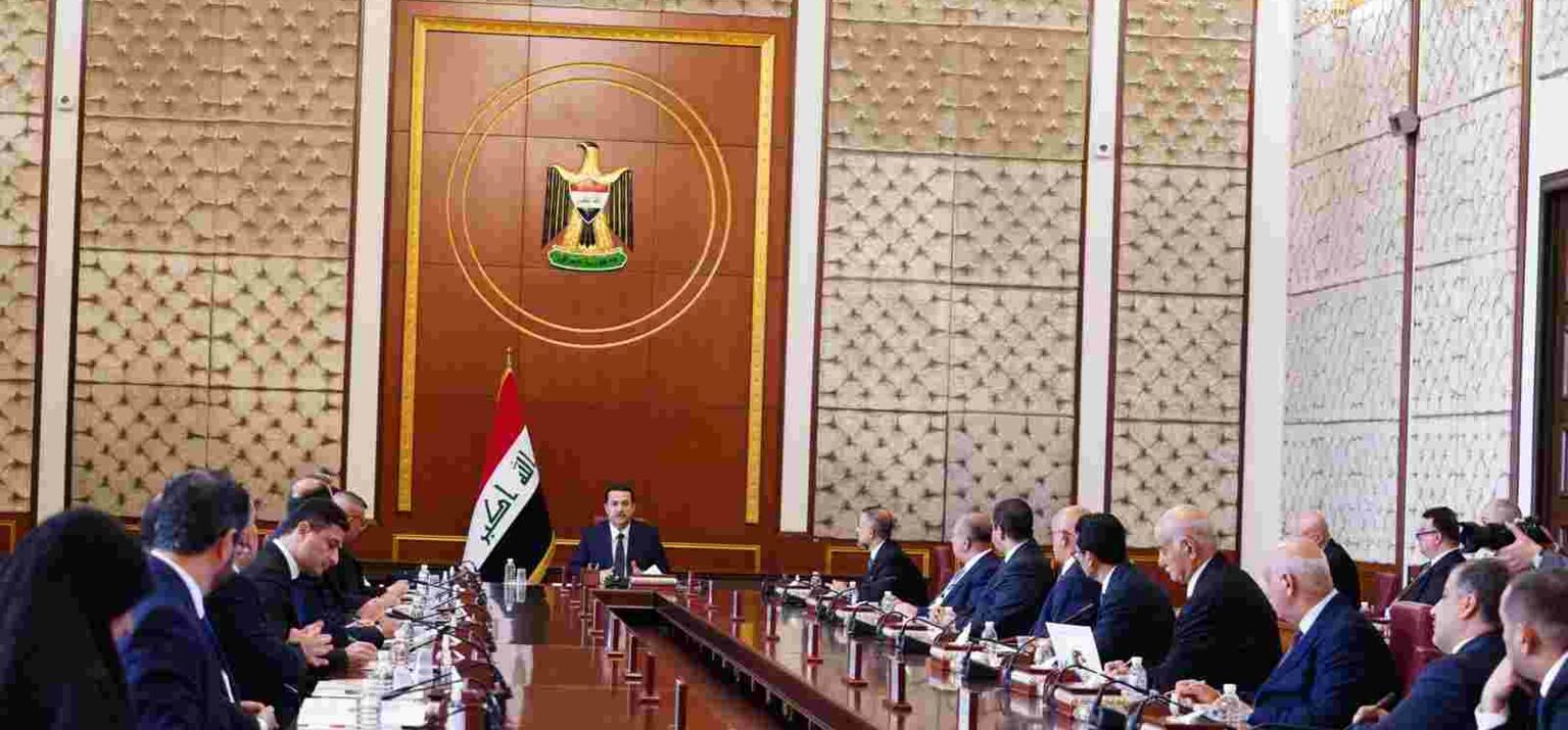 An einem langen Tisch sitzen viele Männer in Anzügen, dem Vorsitzenden, der neben einer irakischen Flagge sitzt, zugewandt
