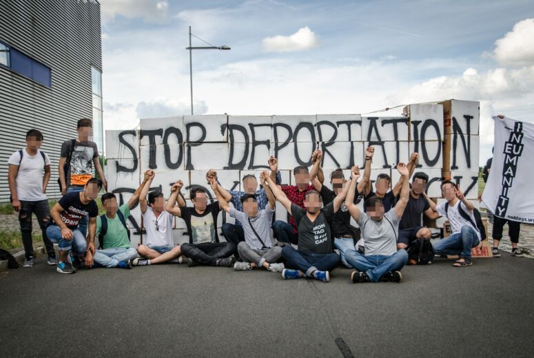 Eine Menschengruppe sitzt auf dem Boden vor einem Plakat mit der Aufschrift »Stop Deportation«