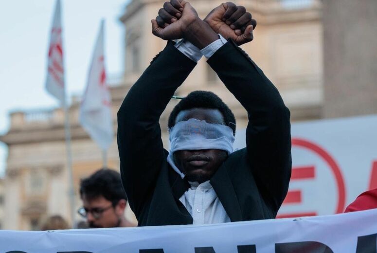 mit verbundene Augen und erhobenen Händen steht Y. David hinter einem Transparent aur der Demonstration gegen italienische Grenzpolitik