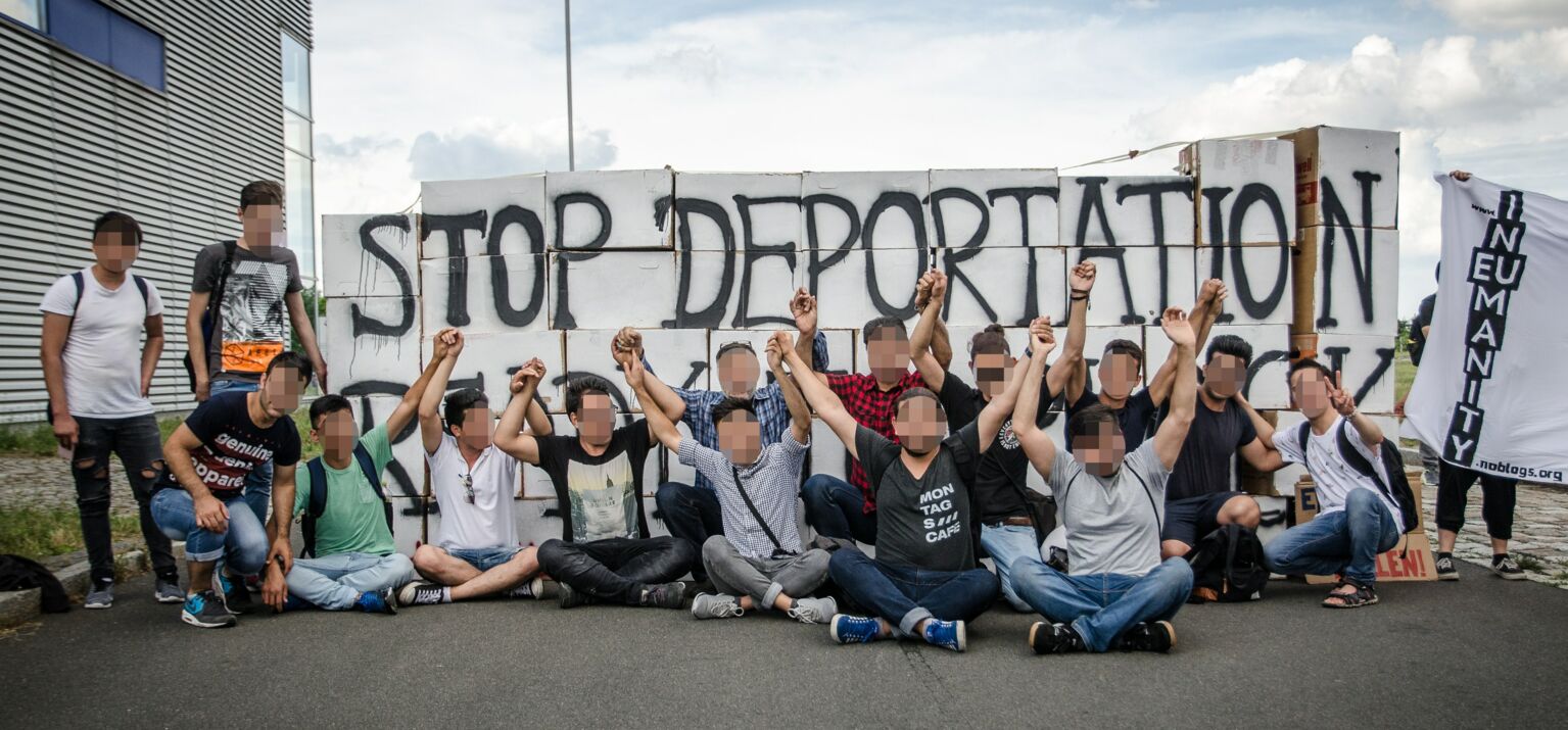 Eine Menschengruppe sitzt auf dem Boden vor einem Plakat mit der Aufschrift »Stop Deportation«