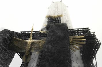 Zu sehen ist ein Teil der Gedenkstätte für die Opfer des Holodomor in Kiew. Ein weißer Turm ragt in den bewölkten Himmel, an dessen unteren Teil eine Skulptur aus messingfarbenen Kranichen und Gittern montiert ist.
