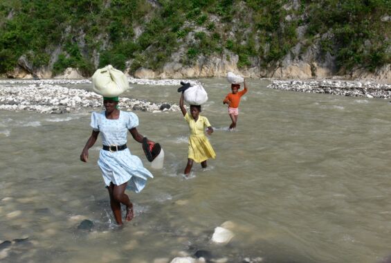 Eine Frau und zwei Kinder mit Büneln auf dem Kopf durchqueren einen Fluss
