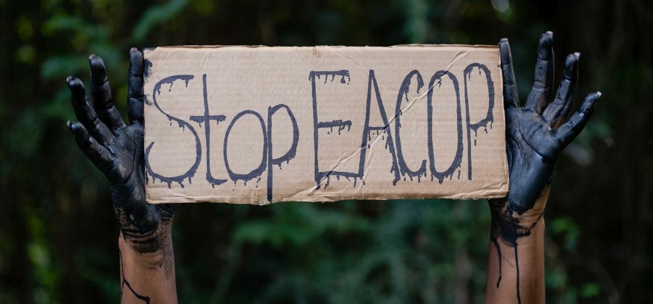 Stop EACOP Kampagne gegen die Rohölpipeline in Uganda und Tansania. Schwarz eingefärbte Hände halten ein Schild mit Stop EACOP hoch.