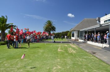 Auf dem Golfplatz Rondebosch Golf Course steht links eine größere Menschengruppe mit roten Fahnen, rechts stehen Golfer*innen unter einem Dach.