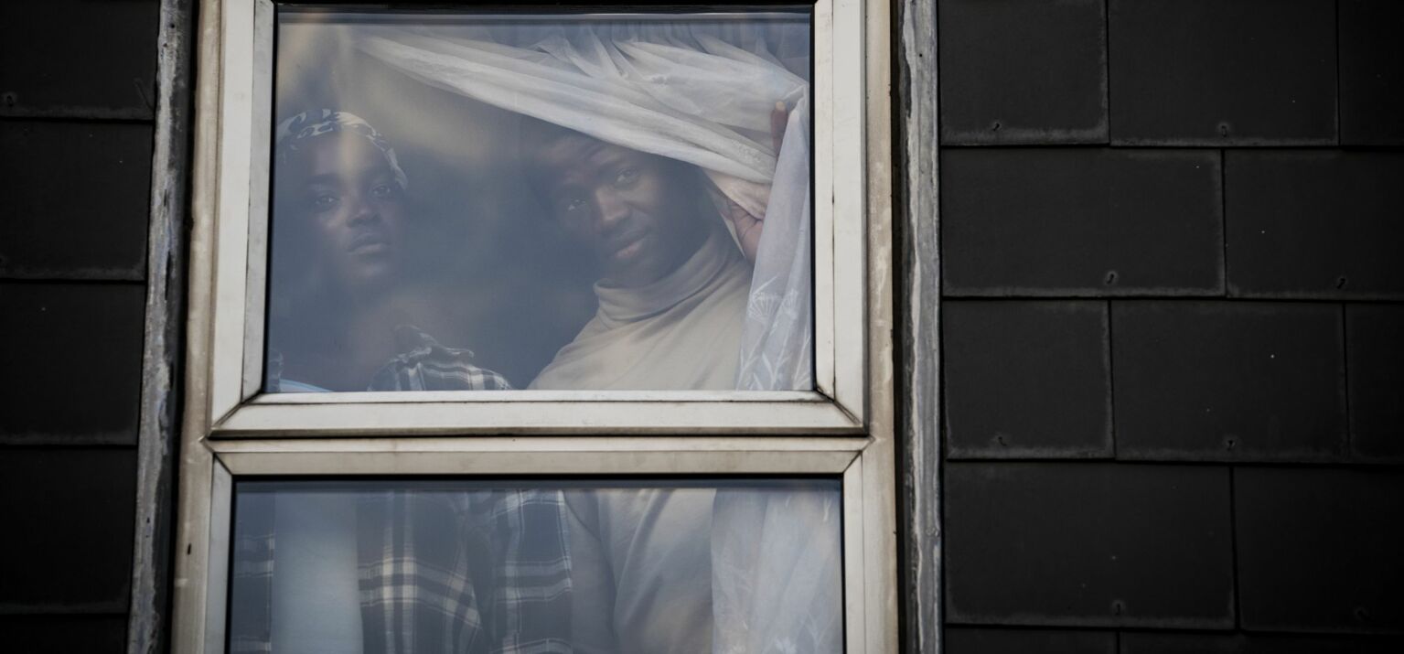 Filmstill »His House« (GB, 2020), Rial (Wunmi Mosaku) und Bol (Sope Dirisu), hinter einem Fenster, Vorhang zur Seite