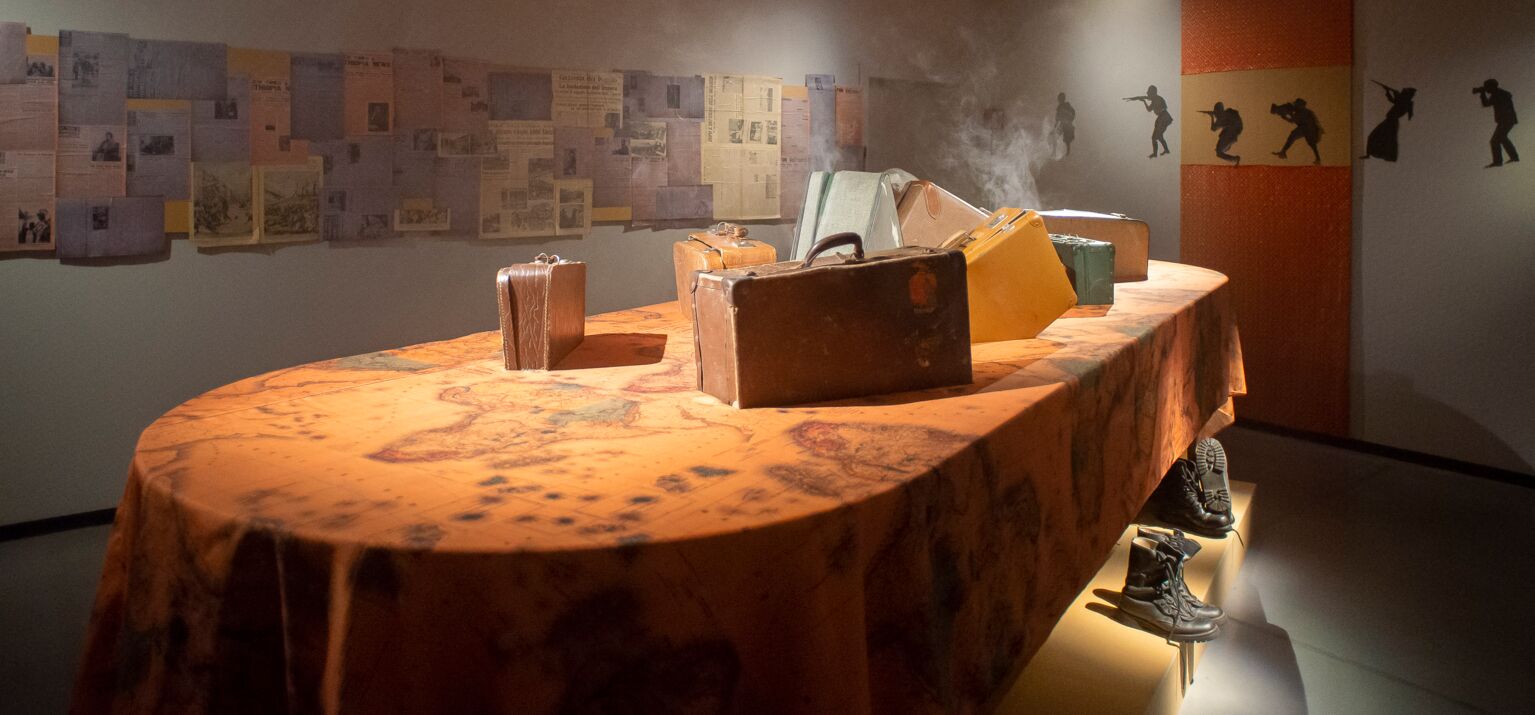 Ein Tisch mit einer Tischdecke mit der Landkarte von Afrika und Koffern darauf steht in einem Ausstellungsraum. Über ihm steigt Rauch auf.