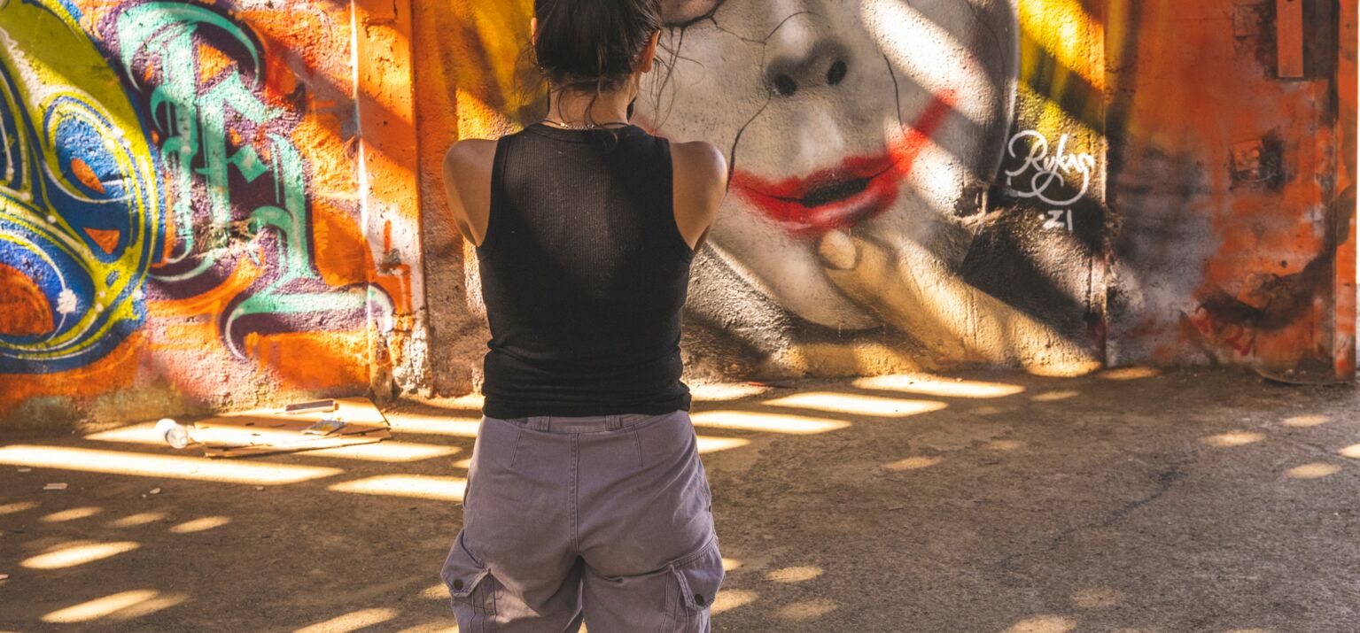 Person, die auf ein Graffit blickt: Eine Maske, die von einem Gesicht gezogen wird. Ist Identität gewählt oder aufgepresst?