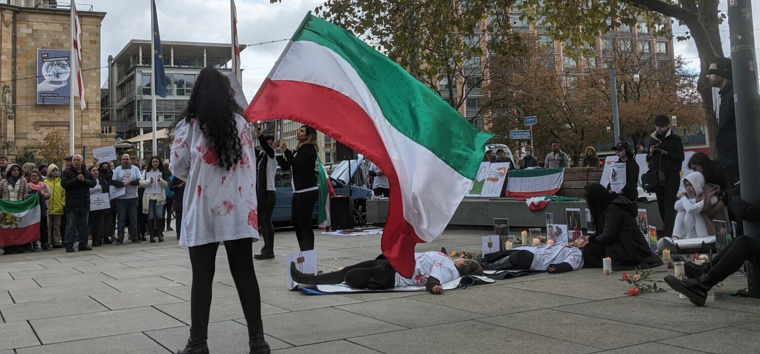 Eine junge Frau mit dunklen Locken und Kunstblzt auf der Kleidung schwenkt eine iranische Flagge