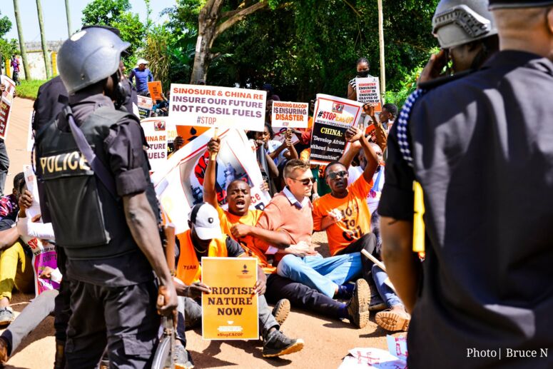 Proteste gegen die EACOP vor der chinesischen Botschaft in Uganda. Protestierende sitzend, umringt von Polizisten, halten Schilder in die Höhe