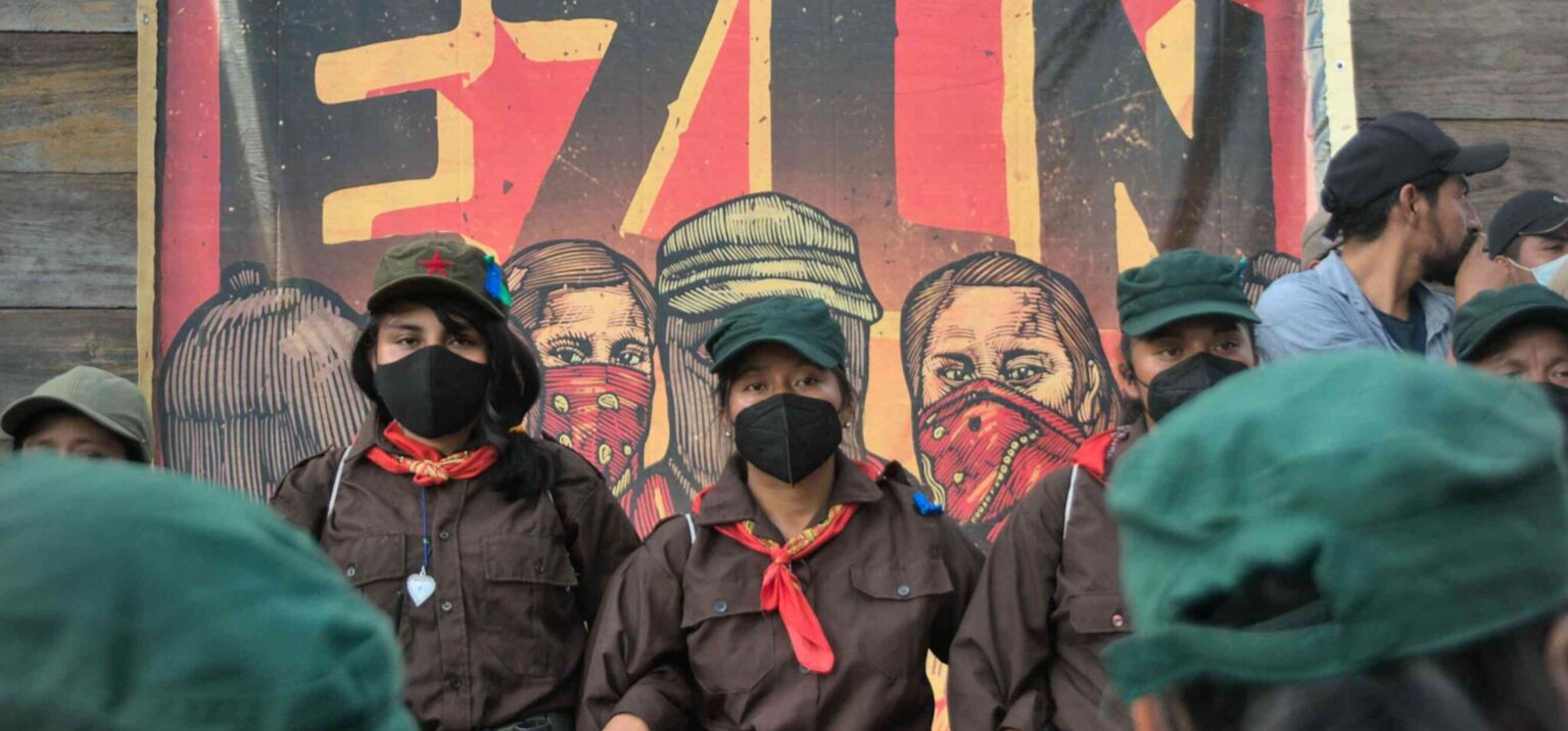 Vermummte EZLN-Kämpfer*innen bei den Jubiläumsfeiern vor einem Plakat in Chiapas