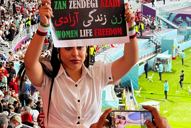 Vor den Rängen eines Fußballstadiums hält eine junge Frau in weißer Bluse ein Schild in die Kamera. Darauf steht in grün-weiß-rot: Zan Zendegi Azadi - Women Life Freedom