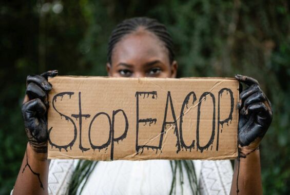 Eine Person mit Erdölbeschmierten Händen hält ein Schild, auf dem steht »Stop EACOP«.