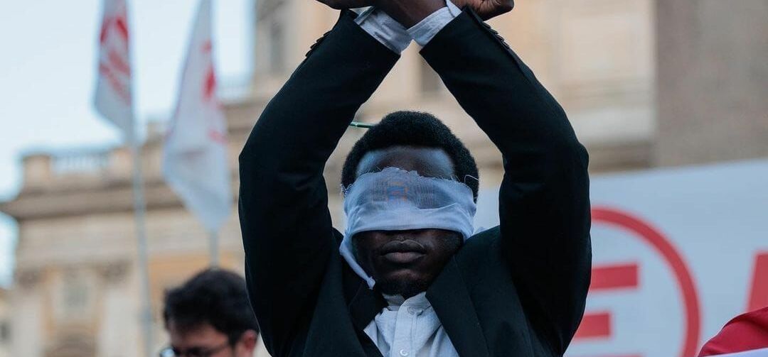 mit verbundene Augen und erhobenen Händen steht Y. David hinter einem Transparent aur der Demonstration gegen italienische Grenzpolitik