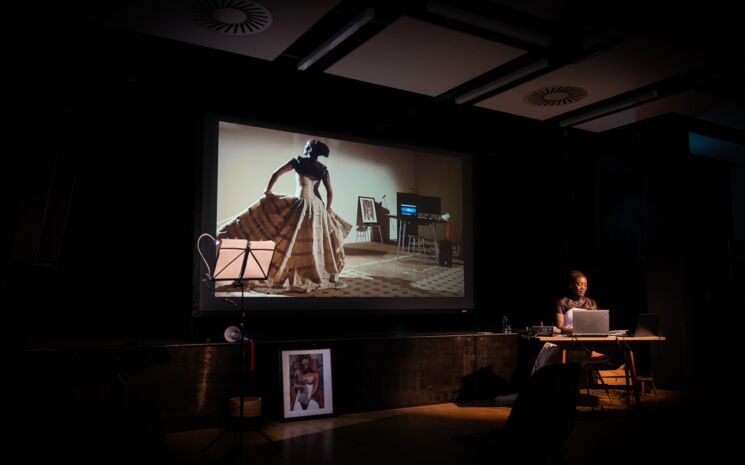 Jasmine Tutum sitzt auf der Bühne, hinter ihr eine Projektion einer Szene. Freiburg, 13. Juni 2020  | Foto: Sévérine Kpoti